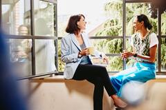 two women having coffee sitting on a windowsill laughing and conversing