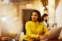 a woman wearing a yellow top smiling while typing on her laptop