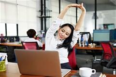 a woman wearing a white long sleeve shirt stretching while in her work cubicle