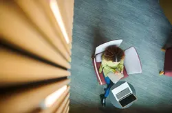 Aerial view of woman writing in a notebook with a computer next to her.