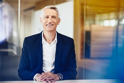 a man wearing a business attire smiling with his arms in front of him placed on a table