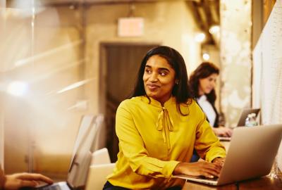 a woman wearing a yellow top smiling while typing on her laptop