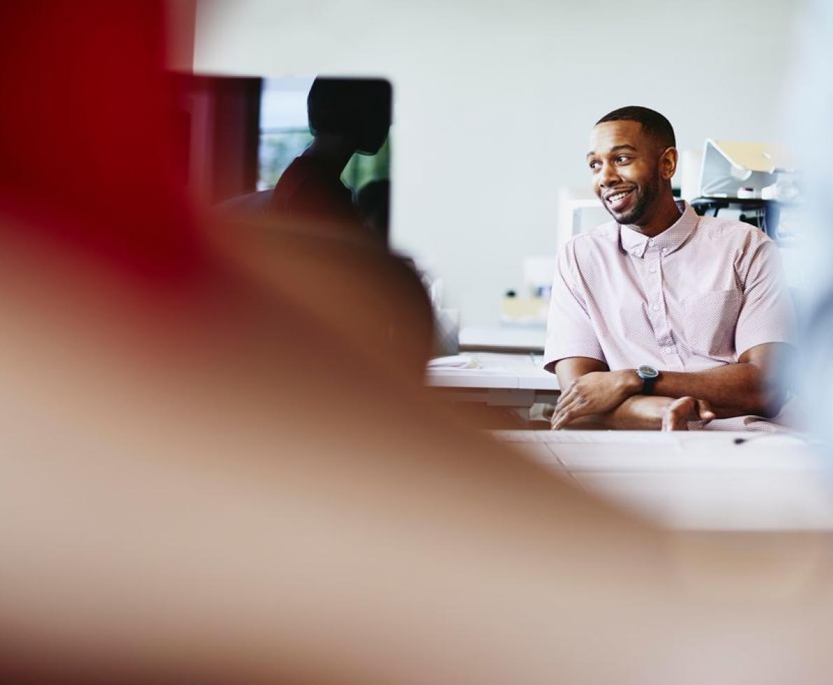 a man talking to a coworker while seated on his office cubicle