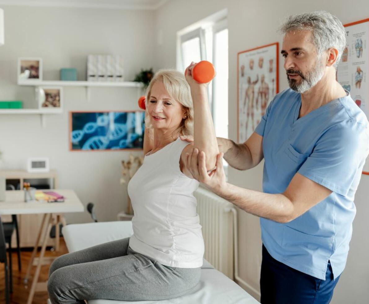 Image of a physiotherapist supporting his patients arm. The patient is holding a weight in each hand.