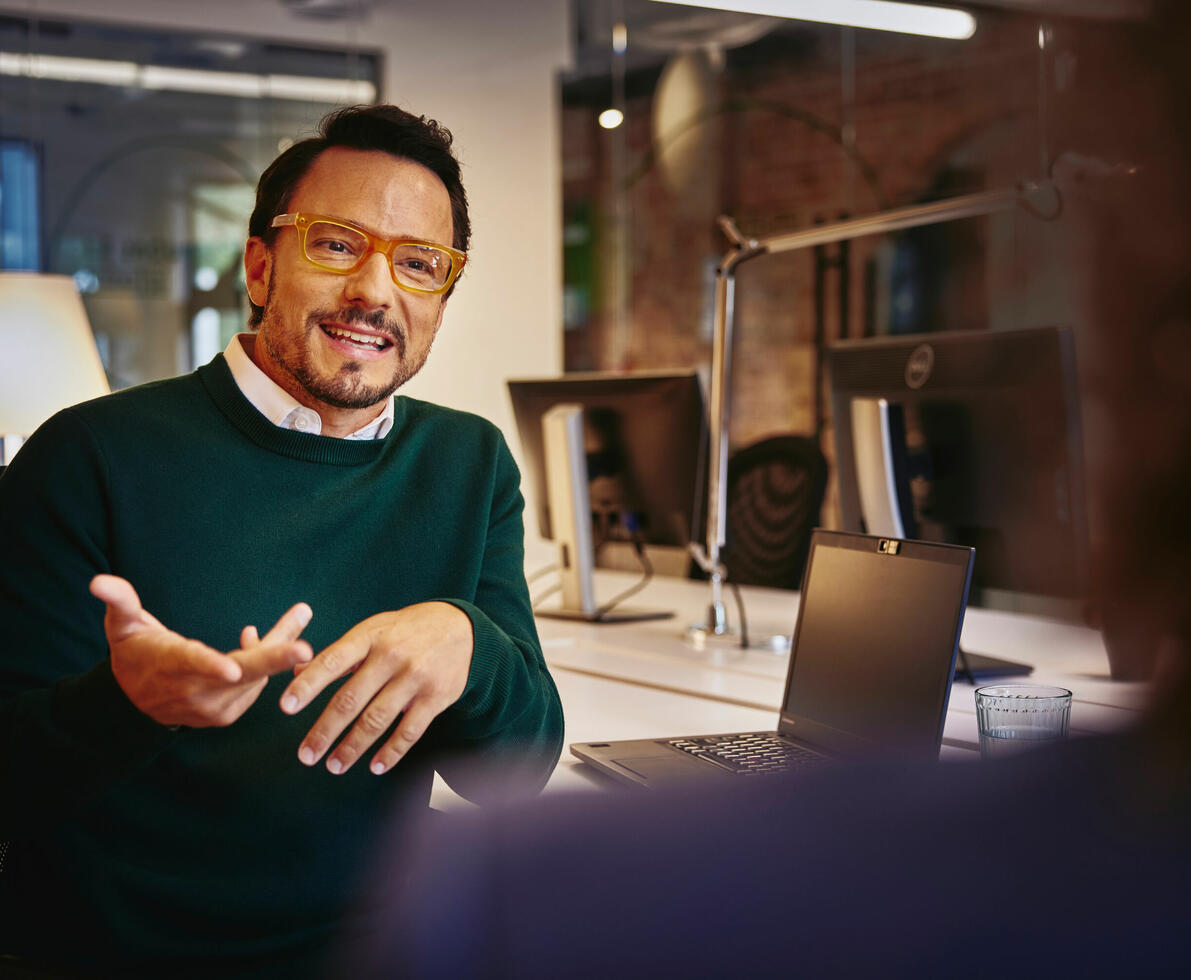 man in green jumper, white shirt underneath, wearing glasses speaking to a colleague in the office