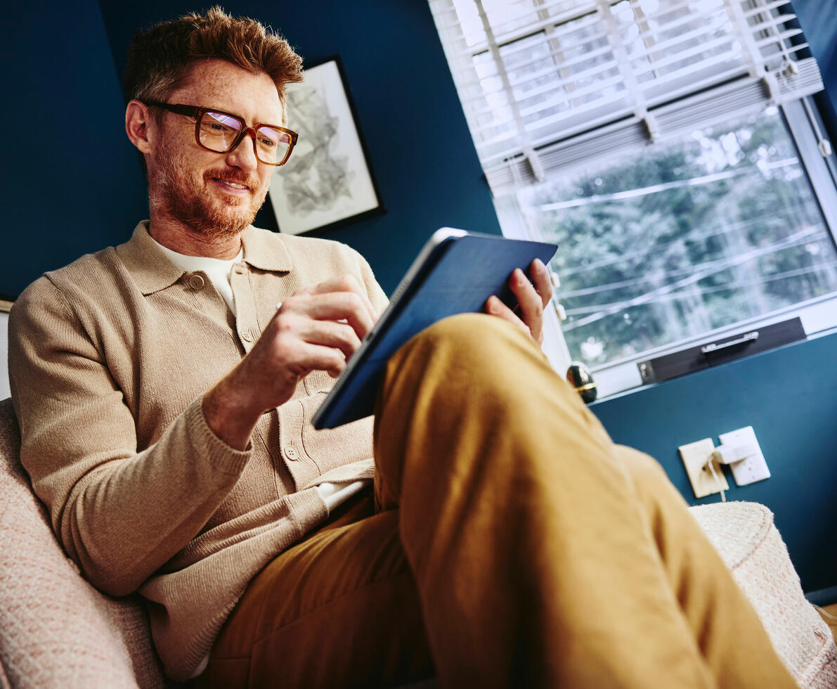 man looking at tablet wearing glasses, legs crossed, wearing brown clothing