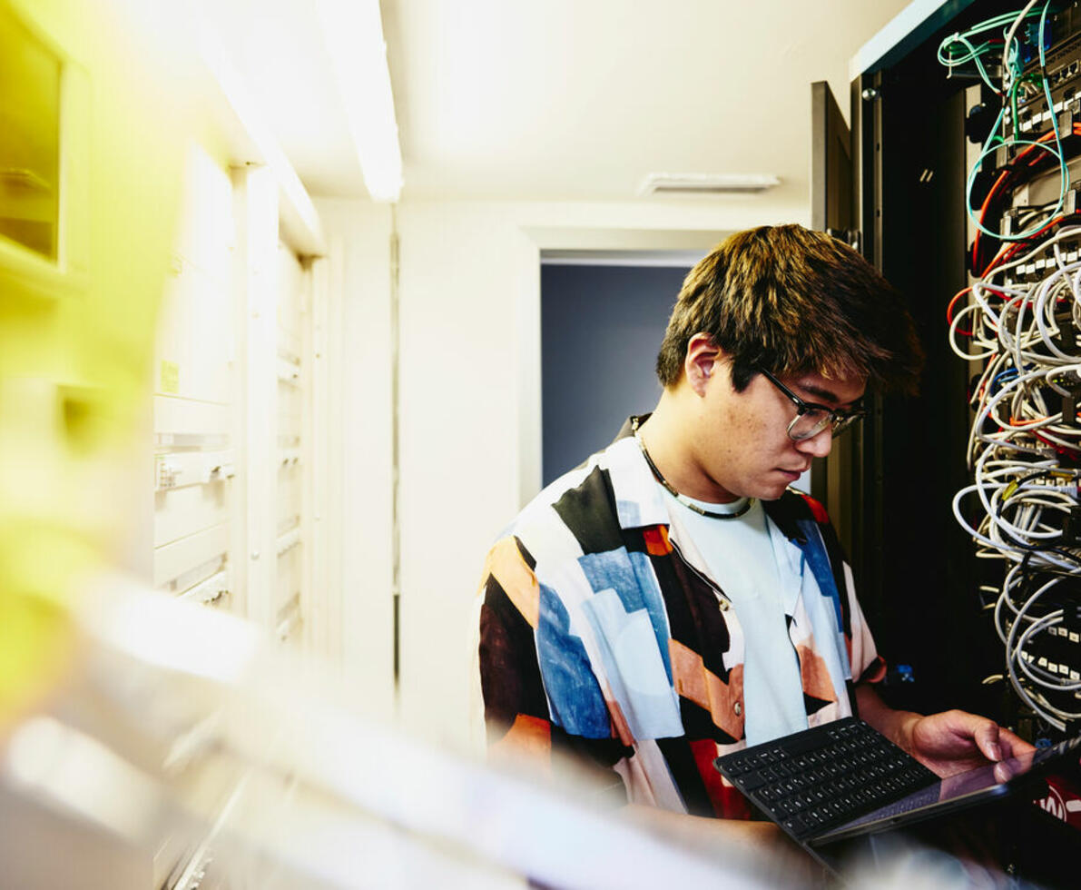 an image of a man with glasses looking at a tablet standing in a servers room.