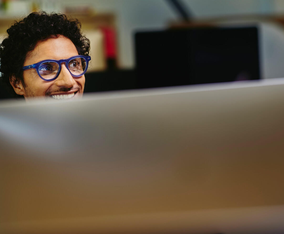 a man wearing glasses peaking from behind his monitor screen while looking to the right