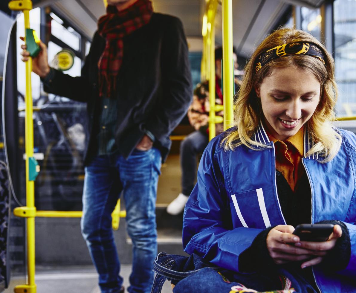 Smiling woman sitting in a bus, looking at her phone. Other people in the background.